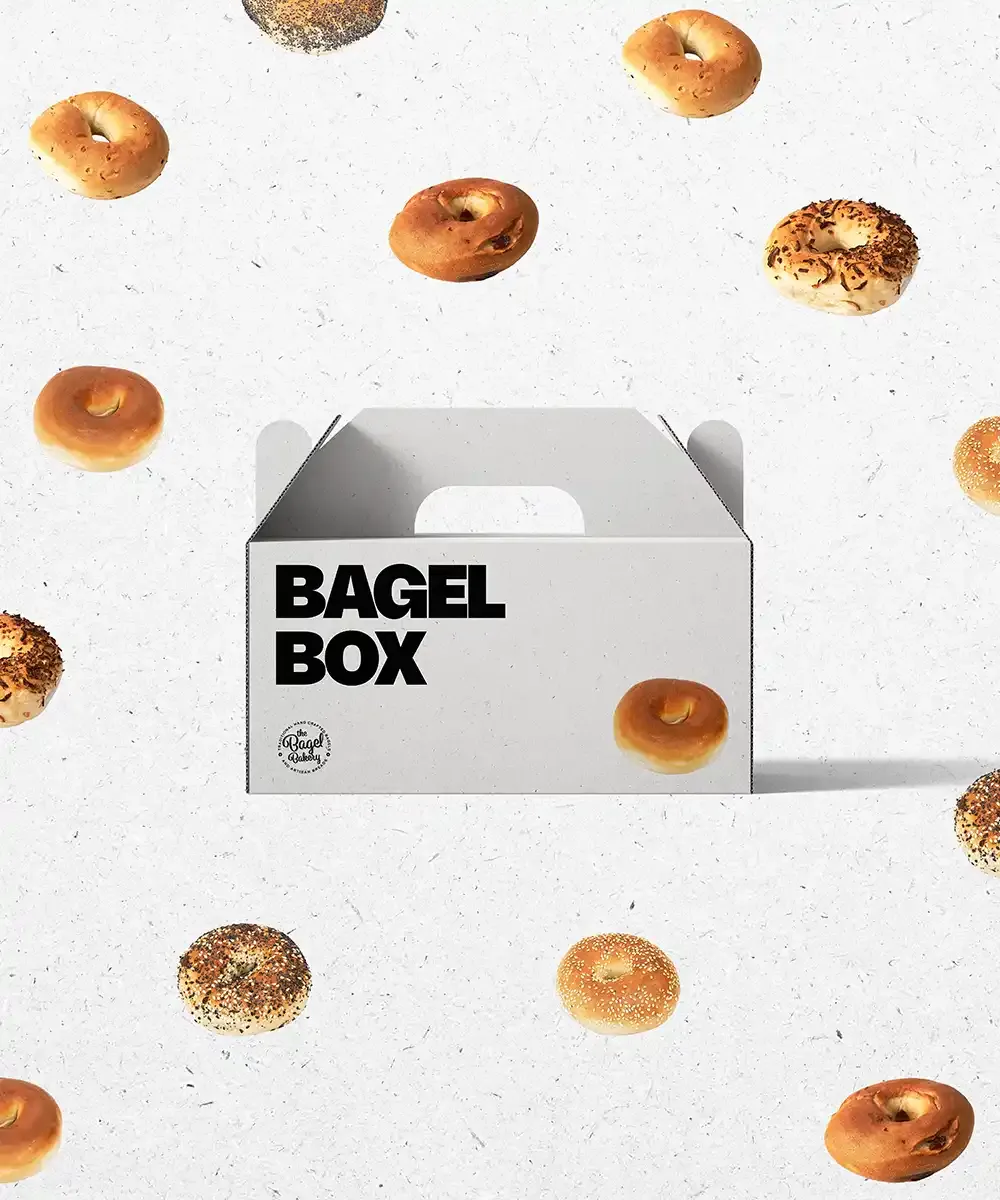 A white Bagel Box gift box with The Bagel Bakery logo, surrounded by floating bagels
